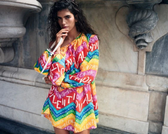 A woman poses in a colorful Valentino dress with a matching purse in front of a marble wall with decorative stonework.