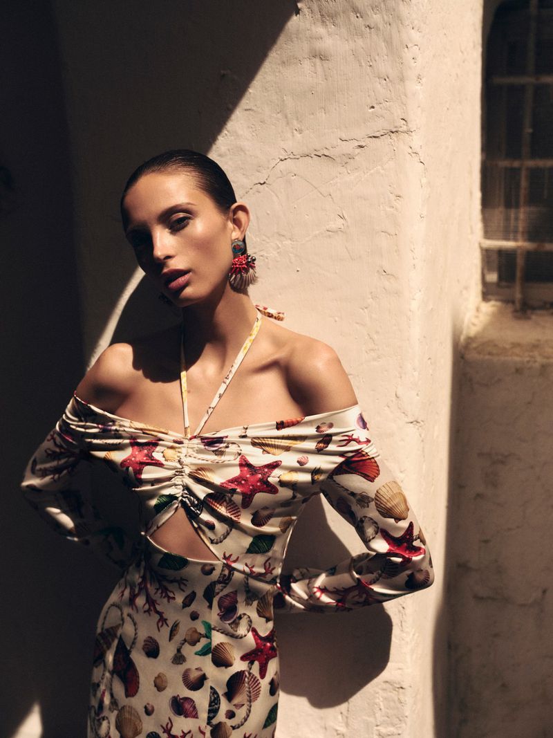 An adult fashion model poses near a whitewashed wall wearing a marine-themed off-shoulder dress with long sleeves and dramatic chandelier earrings.