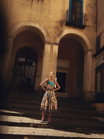 Woman in patterned dress poses on steps with arched architecture in the background. Fashion shot with soft, diffused lighting.