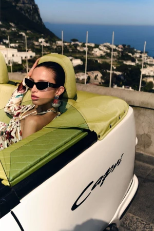 Woman in sunglasses poses in a convertible car with green leather interior, against a coastal city and ocean backdrop on a sunny day.