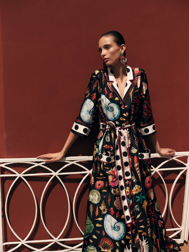 A fashion model showcases a full-length robe with seashells, leaning against white lattice against a red wall, wearing statement earrings.