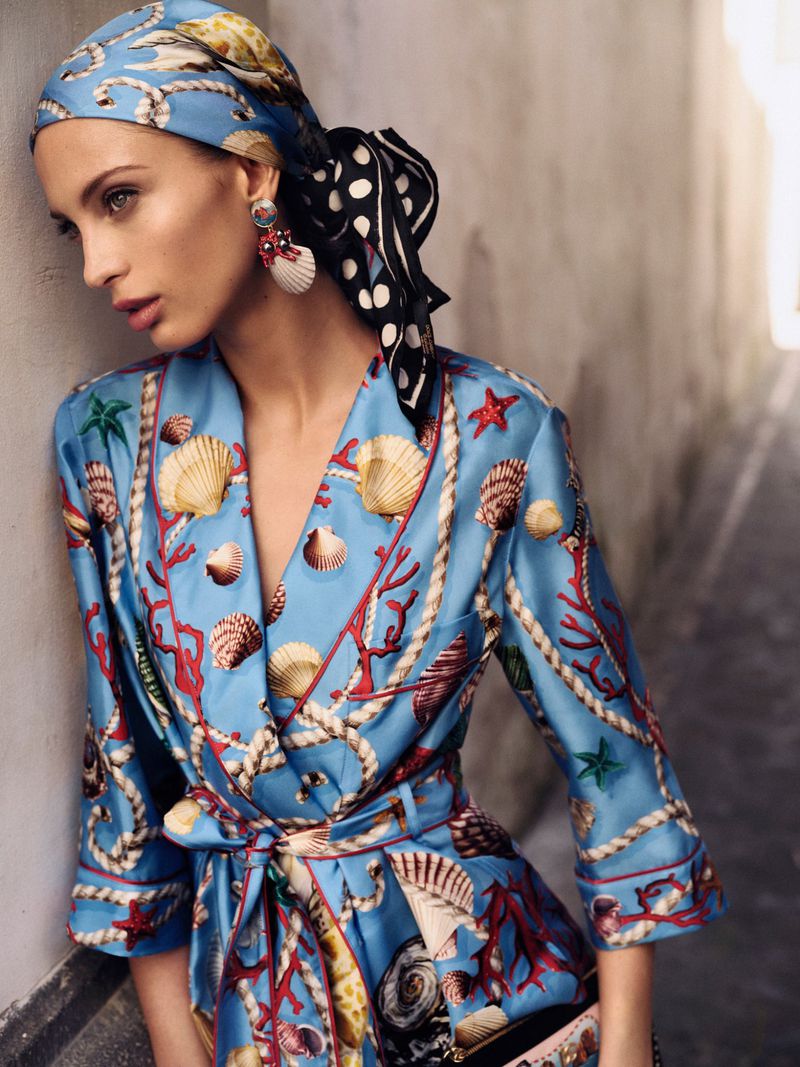 Woman in patterned scarf and robe poses indoors. She has earrings, light skin, and appears to be in her early adult years.