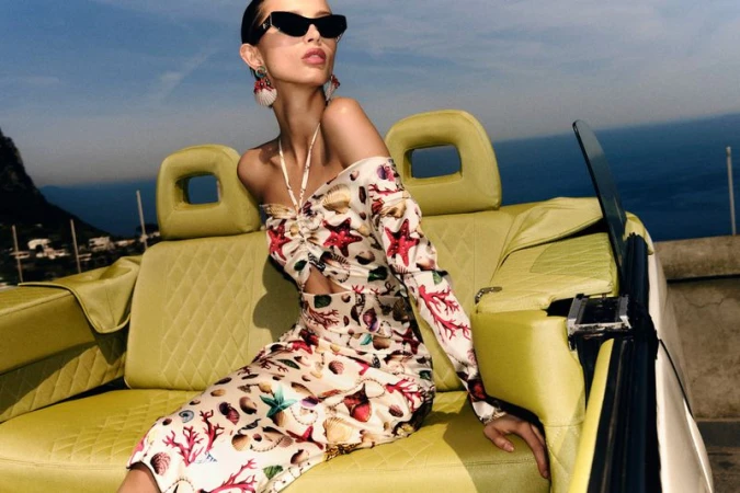 A woman wearing a seashell-patterned dress and sunglasses sits in a green car with a seascape background on a bright day.