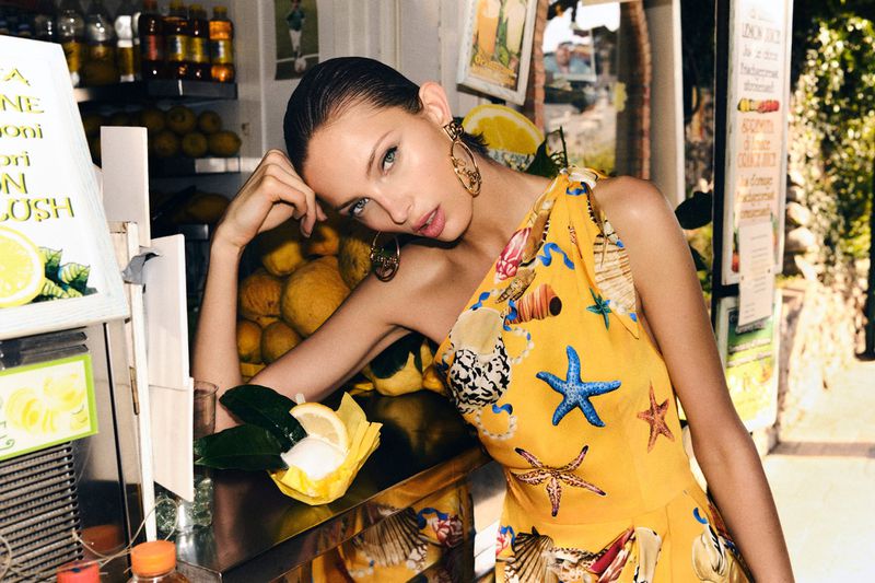 Attractive woman in yellow halter dress with seashell and starfish prints sits near a lemon stand with fresh lemons and juice.
