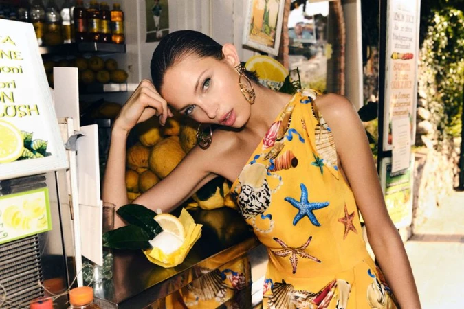 Attractive woman in yellow halter dress with seashell and starfish prints sits near a lemon stand with fresh lemons and juice.