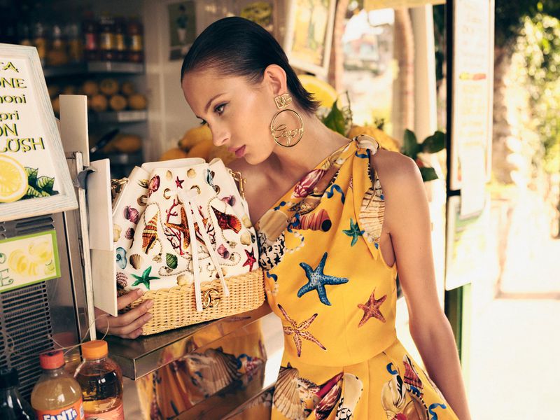 A woman in a yellow seashell-print dress poses with a decorative seashell purse near a beverage stand, showcasing fashion and style.