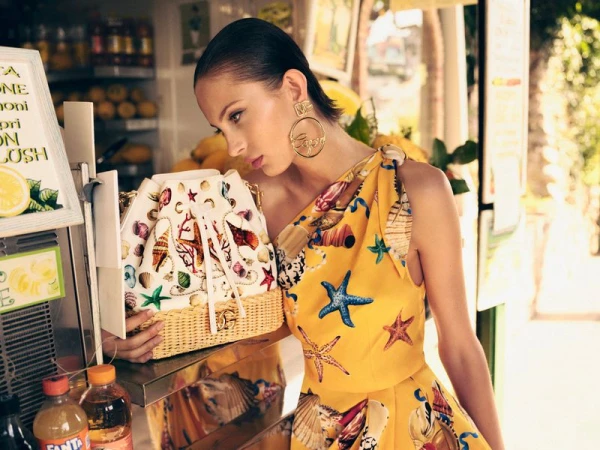 Model Lulu Wood wearing a yellow starfish print dress and large gold Capri earrings holding a seashell pattern straw bucket bag.