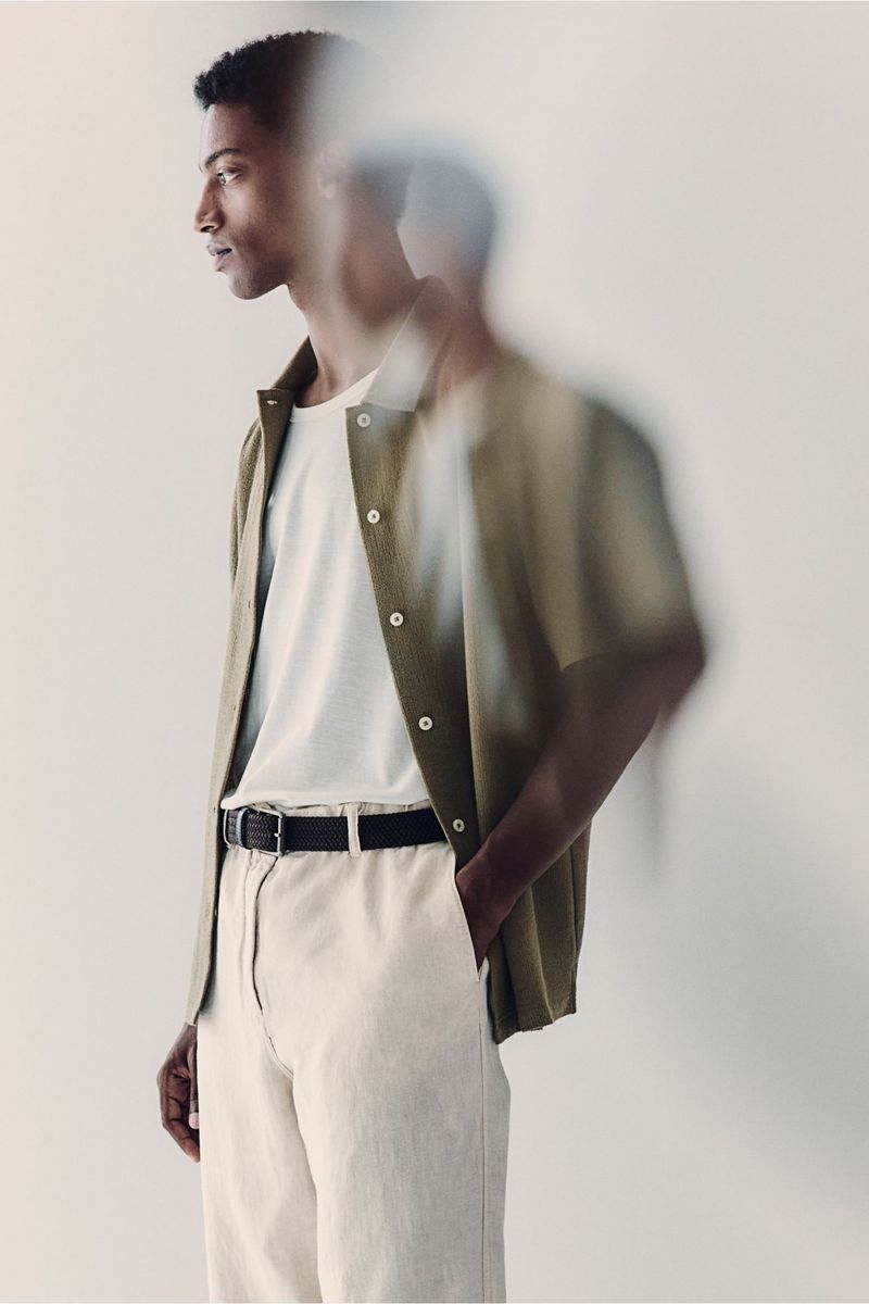 Portrait of a young adult man wearing a short-sleeved button-down shirt with neutral-toned pants, standing indoors against an off-white backdrop.