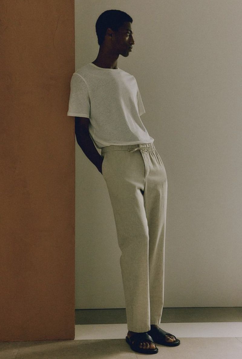 Man stands indoors leaning on wall in relaxed pose wearing a white shirt, tan pants, and black sandals in a fashion editorial.