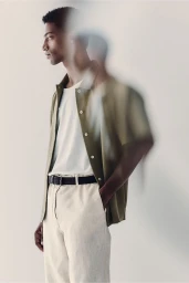 Claudio Goncalves in an olive green short-sleeve shirt, white t-shirt, and cream trousers against a pale studio background.