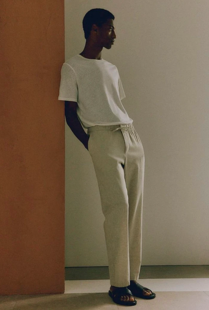 Model Claudio Goncalves leaning against a terracotta partition wearing a white t-shirt and beige drawstring trousers.
