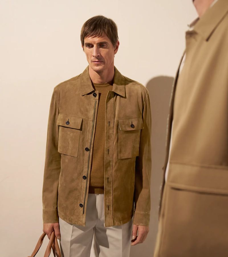 Adult man in a tan suede jacket, brown shirt, and light pants stands against a white background, showcasing a relaxed, stylish look.