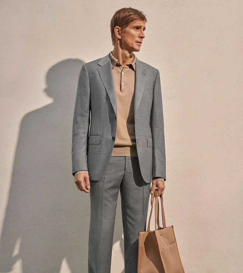 Elegant adult man in a grey suit and beige polo, holding a tote bag against a neutral wall, posing indoors in soft natural light.