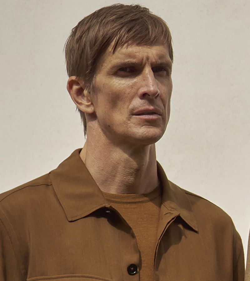 Close-up portrait of a man with light brown hair, wearing a brown shirt, against an off-white background. Serious expression.