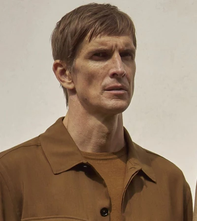Close-up portrait of a man with light brown hair, wearing a brown shirt, against an off-white background. Serious expression.