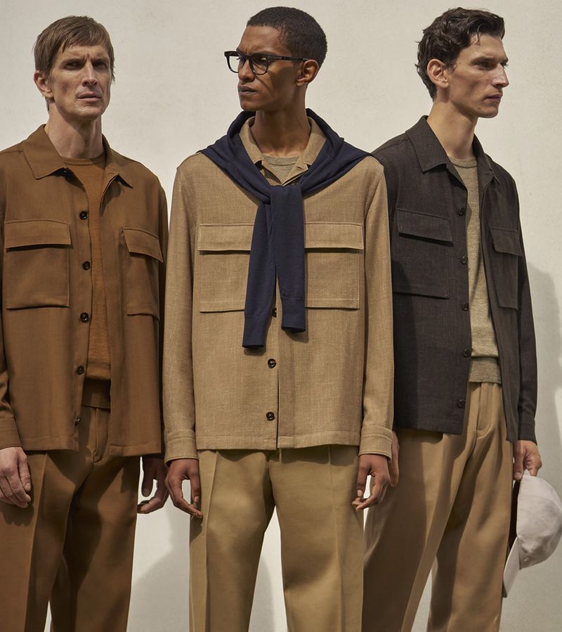 Three adult males pose indoors wearing casual tan and brown collared button-down shirts with matching pants in a fashion setting.