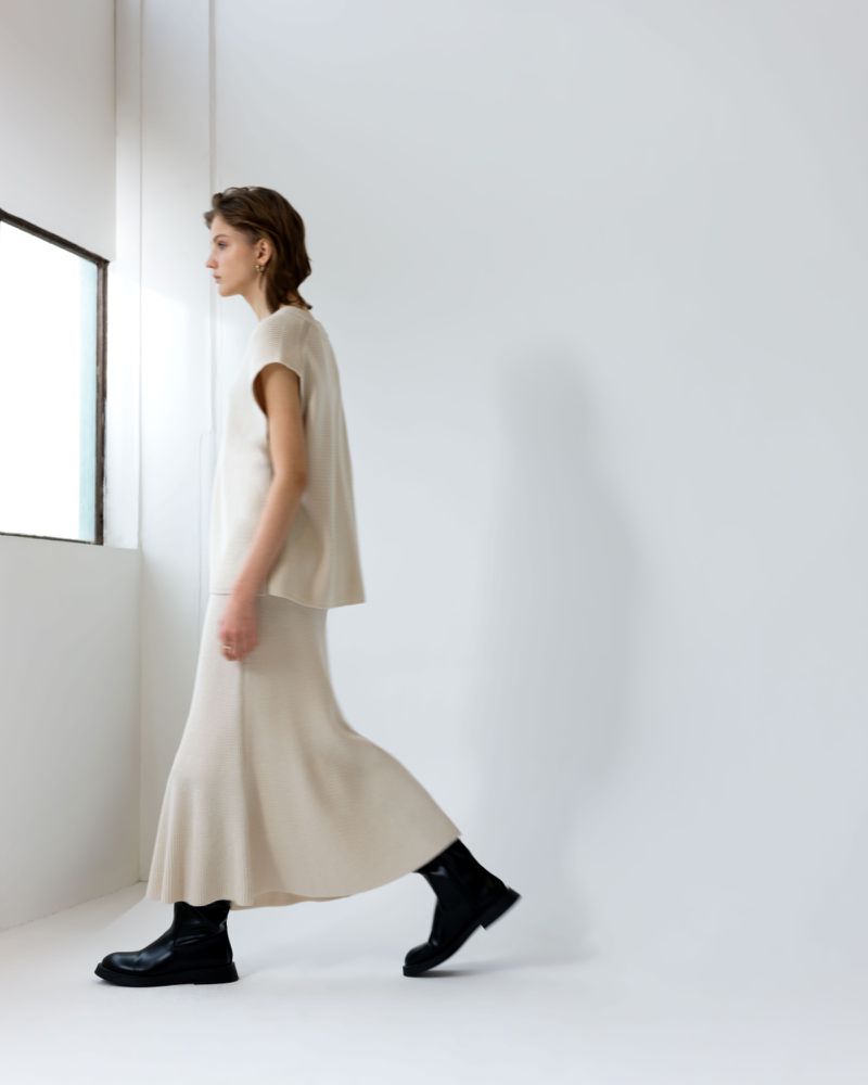 A woman in a beige two-piece outfit and black boots walks against a stark white wall with a window on the left side of the frame.