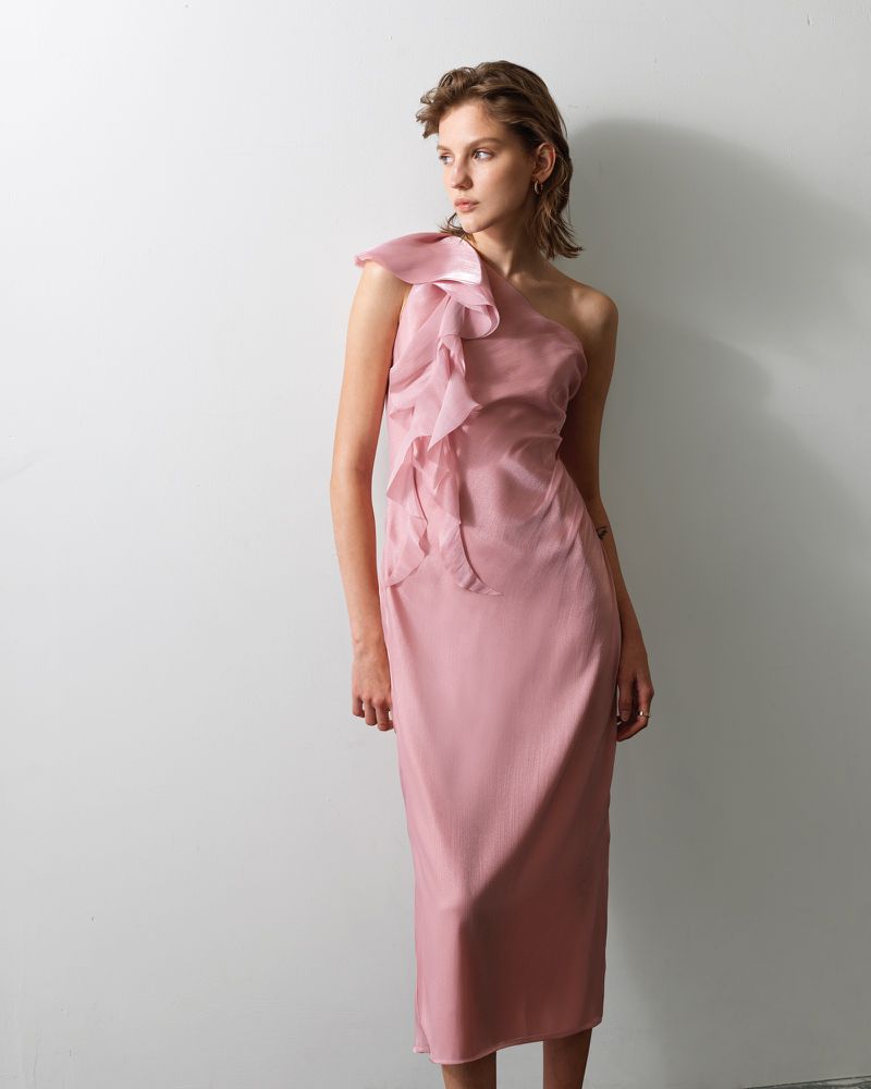 Elegant young woman in a light pink, one-shoulder dress with ruffle detail, standing against a simple white background in a studio setting.
