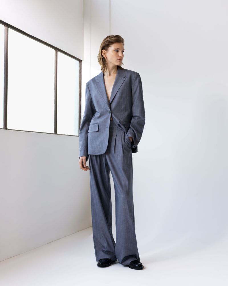 A slender person poses in a relaxed stance indoors, sporting a fashionable grey-blue suit with wide-leg pants and polished black shoes.