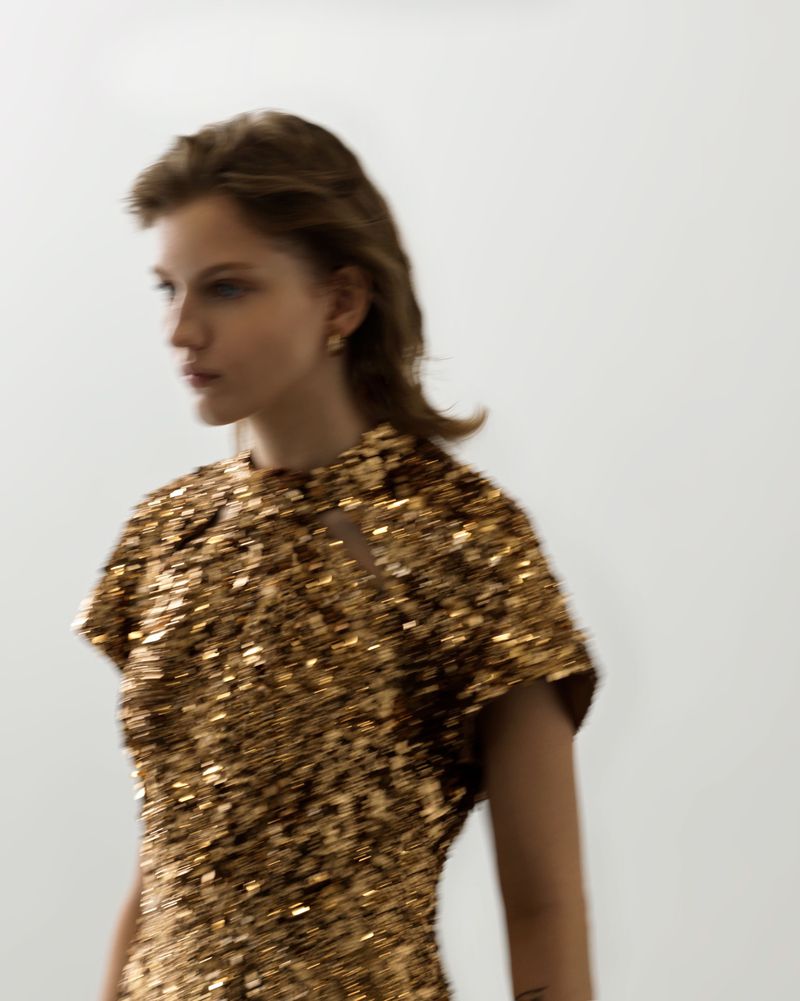 Portrait of a fair-skinned young adult female with light brown hair wearing a gold sequin dress, looking left in a blurry, brightly lit photo studio.