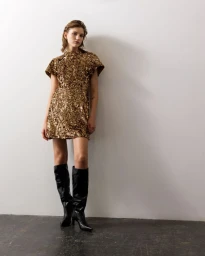 Model Nadine Kirilova posing in a metallic gold sequin mini dress and knee-high black leather boots against a white studio wall.