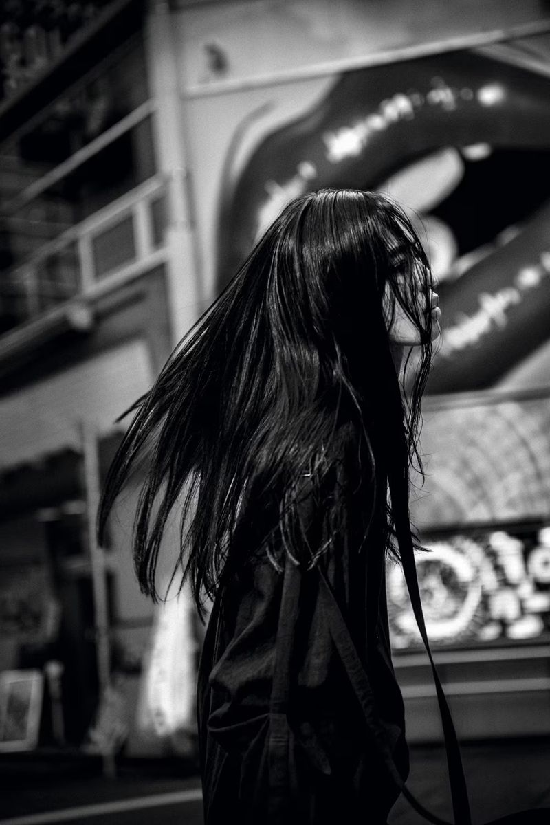 Monochrome portrait of an East Asian woman with long dark hair blowing in the wind against a building facade with a large lips poster.