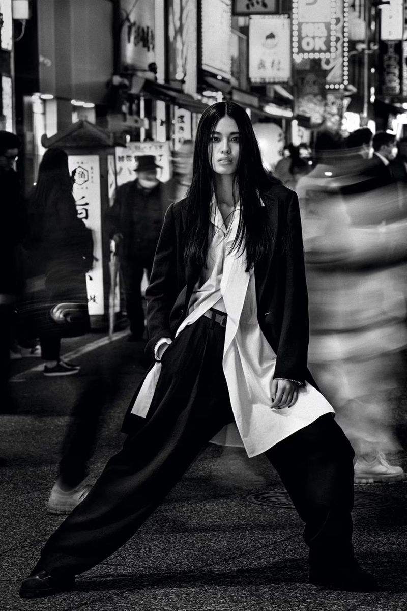 Fashionable woman strikes a confident pose on an urban street at night, wearing a suit with wide-leg pants against a backdrop of blurred pedestrians.