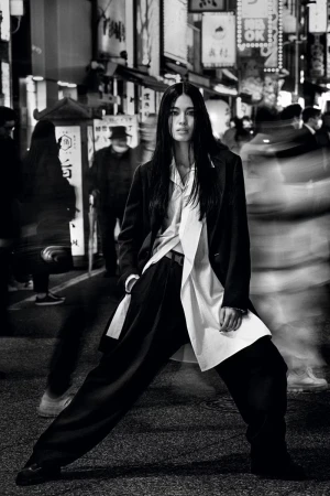 Fashionable woman strikes a confident pose on an urban street at night, wearing a suit with wide-leg pants against a backdrop of blurred pedestrians.
