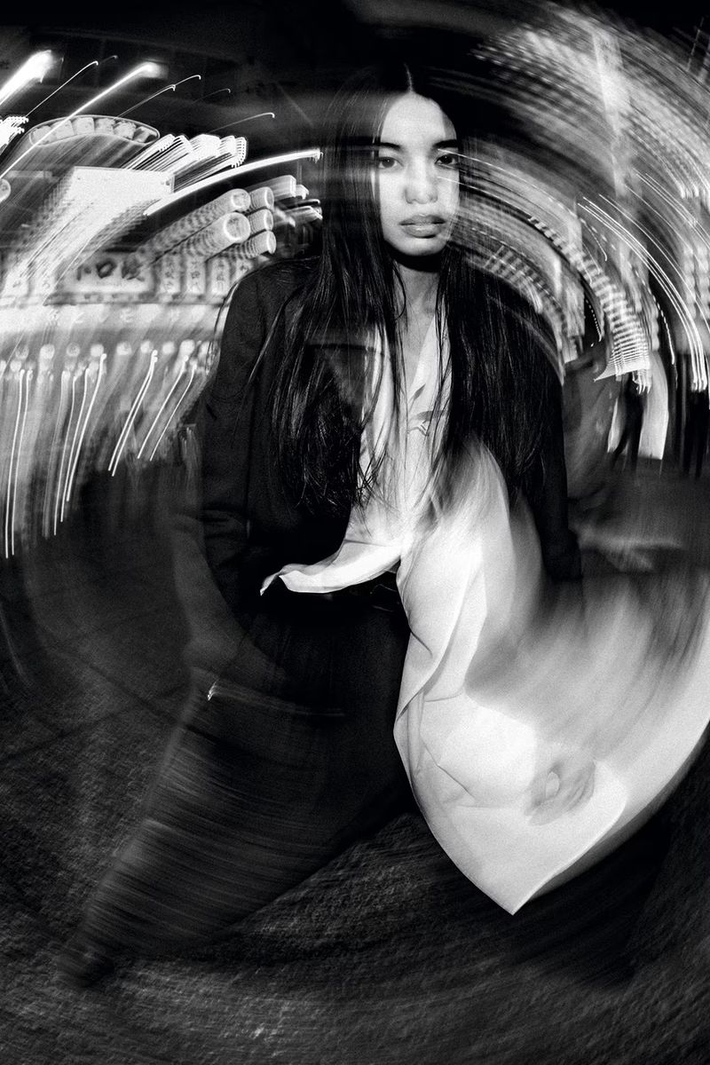 Portrait of a long-haired woman wearing a jacket, standing outdoors at night with blurred city lights creating a dynamic motion effect.