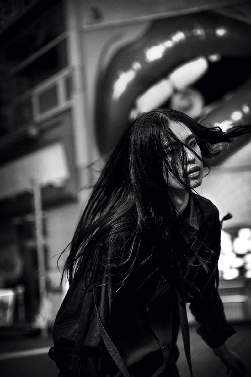 Monochrome portrait of a young adult with long black hair posing in front of a building facade with large lips sculpture.