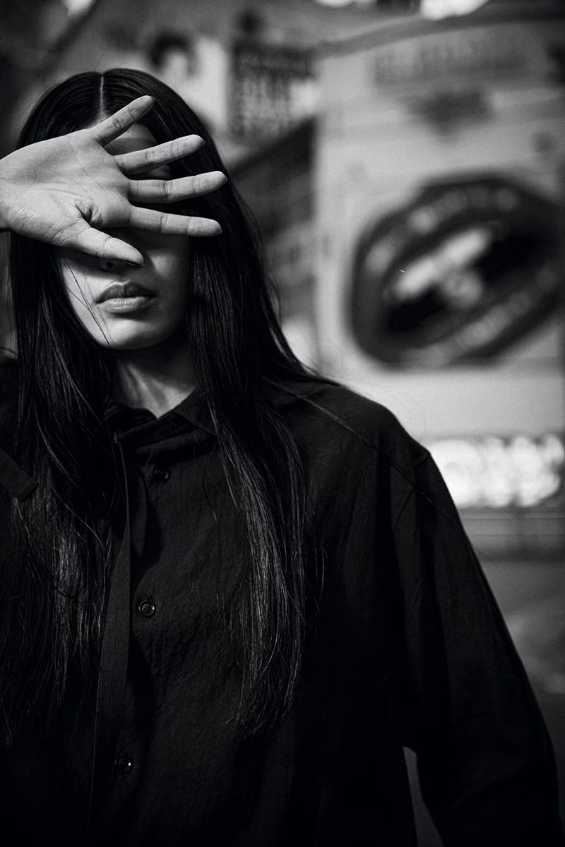 Portrait of a young woman with long dark hair wearing a black button-down shirt and covering her face with her hand indoors.