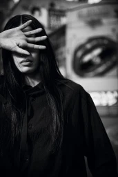 Black and white portrait of a woman with long dark hair holding her hand up to block her eyes against an urban street background.