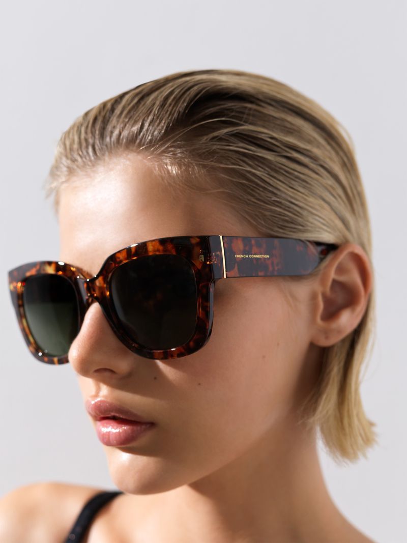 Blonde woman models tortoiseshell sunglasses with green lenses indoors against a neutral background in a fashionable portrait shot.