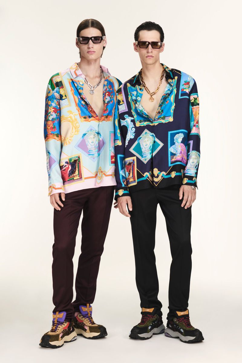 Two adult men pose in patterned shirts, dark trousers, and chunky shoes against a bright backdrop, accessorized with sunglasses and necklaces.
