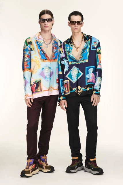 Two adult men pose in patterned shirts, dark trousers, and chunky shoes against a bright backdrop, accessorized with sunglasses and necklaces.