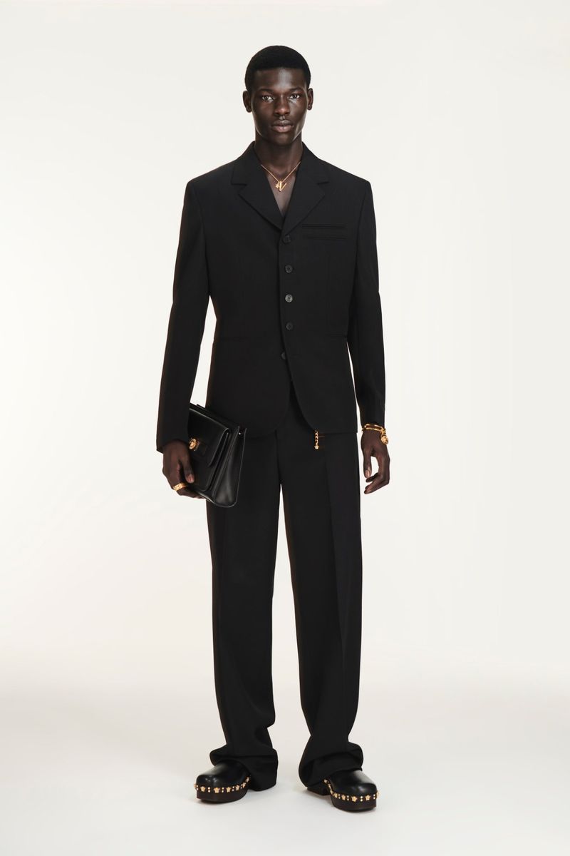 A Black male model showcases a black suit, holding a purse in a full shot against a white backdrop, accessorized with gold jewelry.