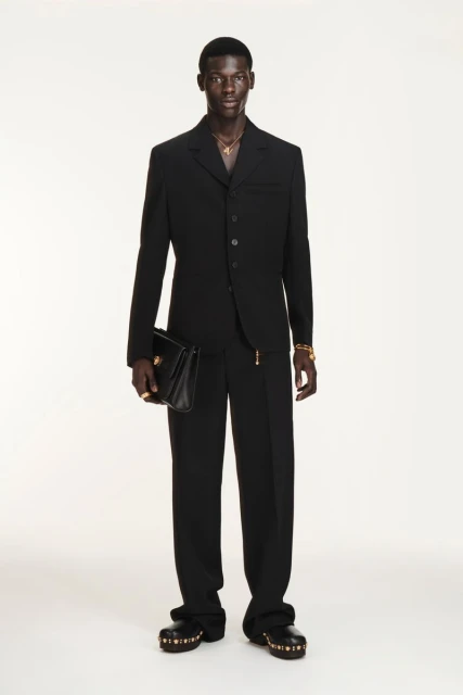 A Black male model showcases a black suit, holding a purse in a full shot against a white backdrop, accessorized with gold jewelry.