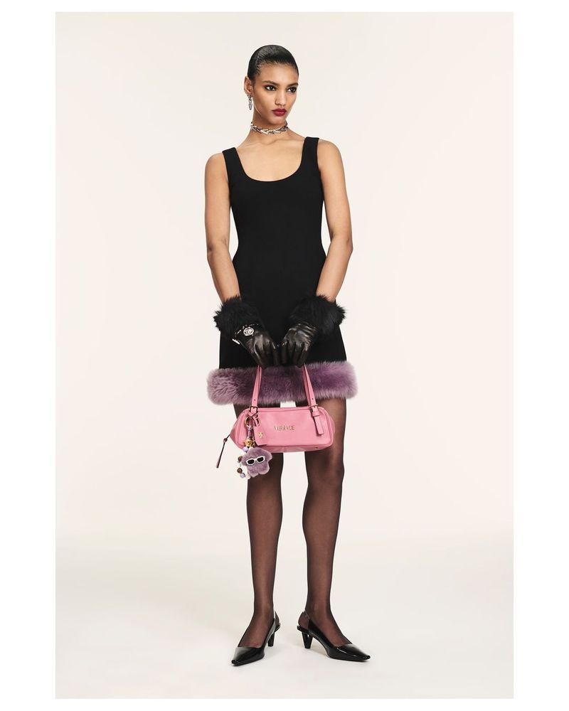 Full shot of a slender woman in a black dress, stockings, and heels, holding a pink purse with a furry charm, set against a plain backdrop.