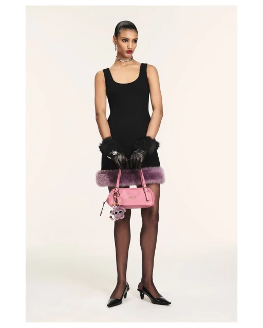 Full shot of a slender woman in a black dress, stockings, and heels, holding a pink purse with a furry charm, set against a plain backdrop.
