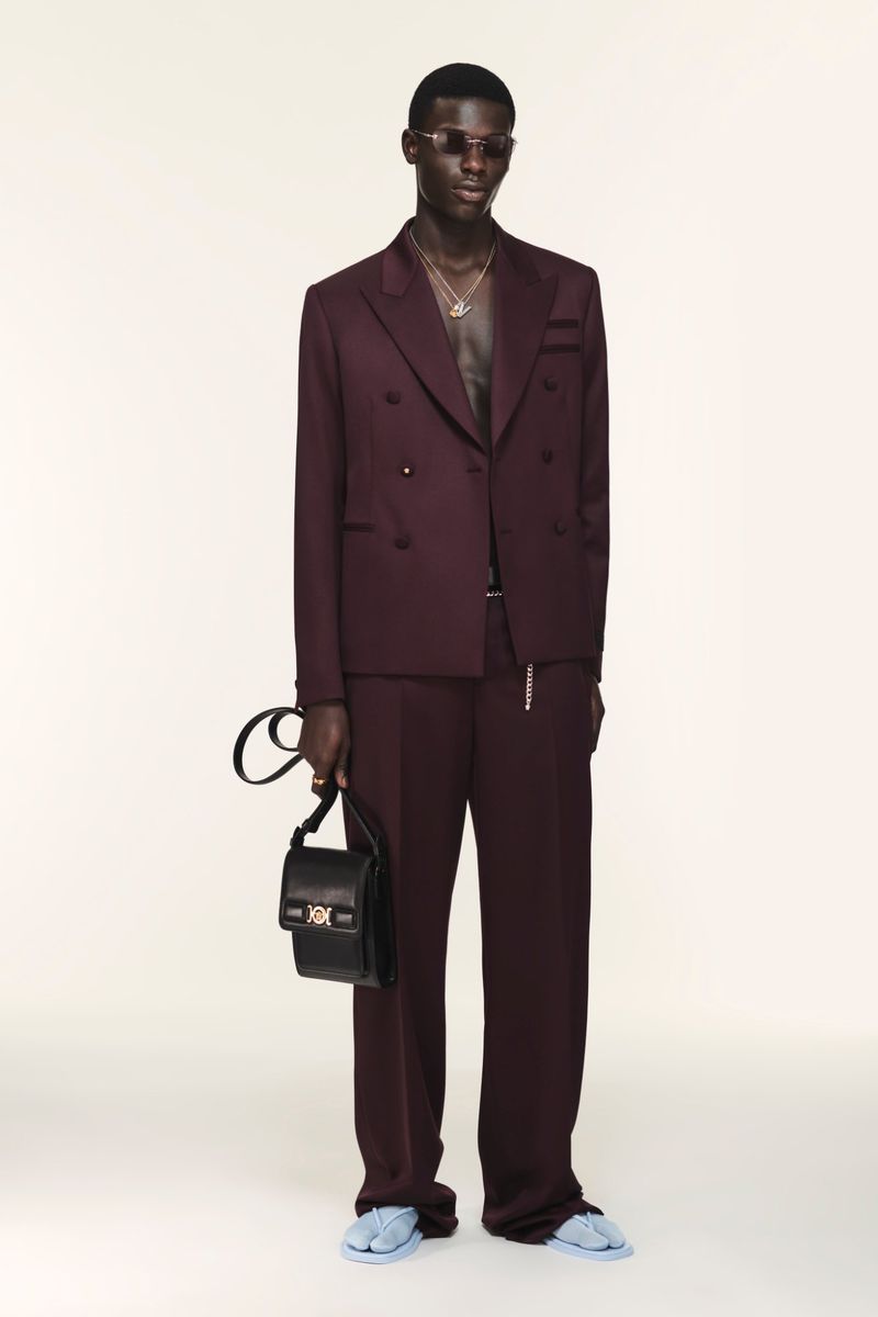 Man poses in a plum double-breasted suit with a black bag against a white backdrop in a fashion studio shot for a luxury brand.