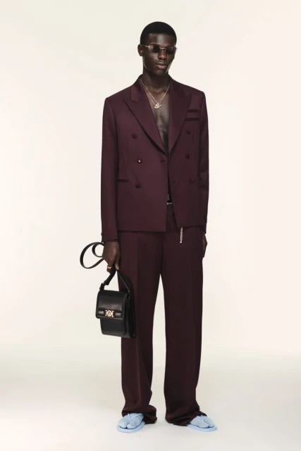 Man poses in a plum double-breasted suit with a black bag against a white backdrop in a fashion studio shot for a luxury brand.