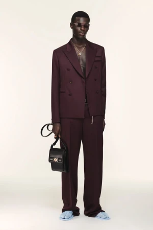 Man poses in a plum double-breasted suit with a black bag against a white backdrop in a fashion studio shot for a luxury brand.