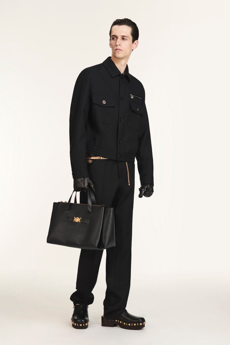 A slender male model in a black jacket, trousers, and shoes holds a black tote bag in a studio setting on a white background.