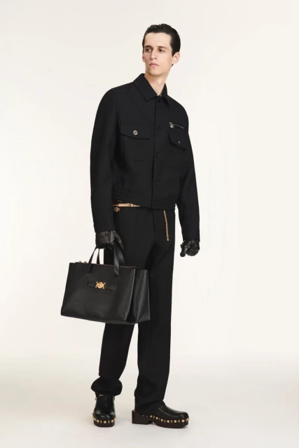 A slender male model in a black jacket, trousers, and shoes holds a black tote bag in a studio setting on a white background.