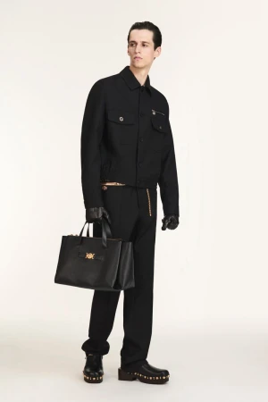 A slender male model in a black jacket, trousers, and shoes holds a black tote bag in a studio setting on a white background.