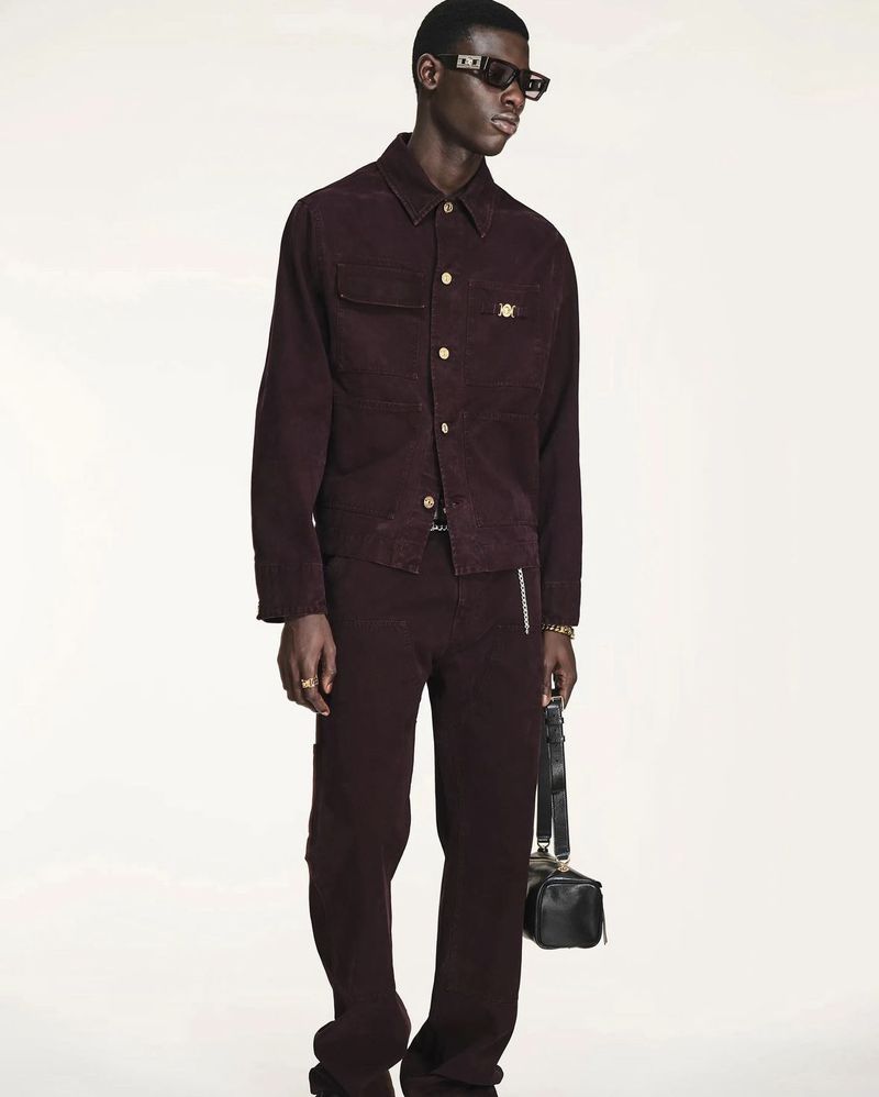 A tall, dark-skinned man is showcased in a full-body shot, wearing a maroon corduroy jacket, matching pants, and black sunglasses against a white backdrop.