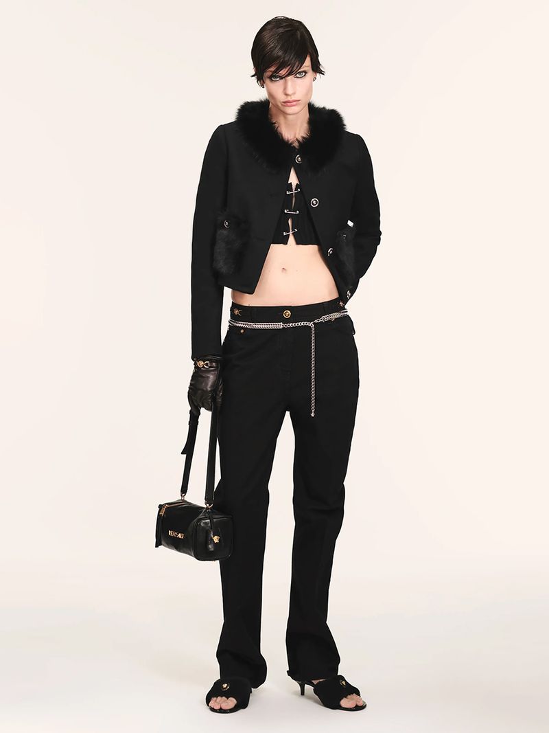 Fashion model in a black cropped jacket with fur trim, black pants, and open-toe heels poses against a white background in a studio shot.