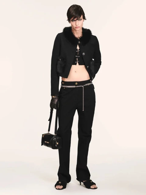 Fashion model in a black cropped jacket with fur trim, black pants, and open-toe heels poses against a white background in a studio shot.