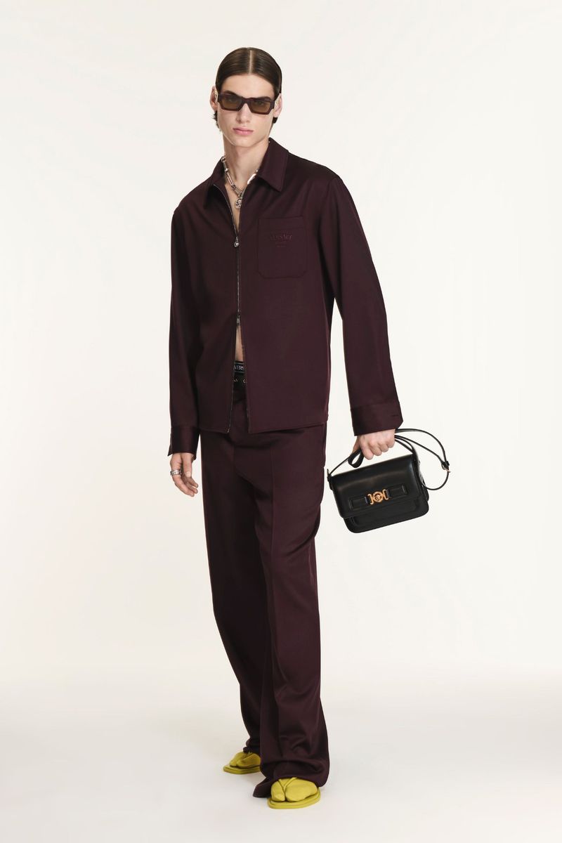 A slender adult male model poses indoors wearing a maroon collared shirt, maroon pants, sunglasses, jewelry, and yellow sandals with a black purse.