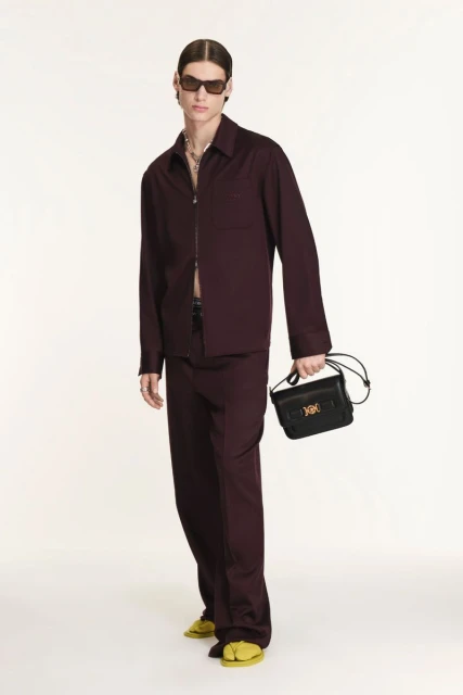 A slender adult male model poses indoors wearing a maroon collared shirt, maroon pants, sunglasses, jewelry, and yellow sandals with a black purse.